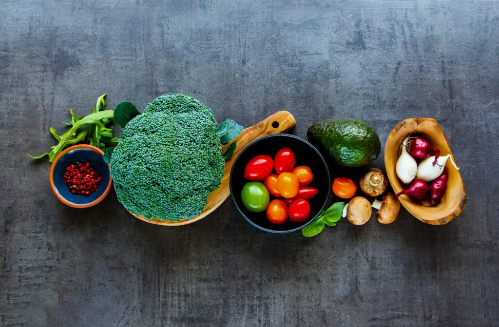 Fresh vegetables including broccoli, tomatoes, avocado, onions, mushrooms, and greens arranged on a dark surface.