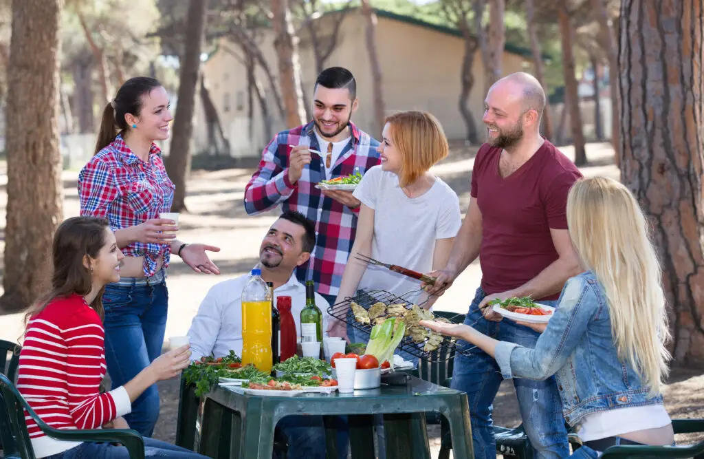 Group of friends enjoying a barbecue picnic in a park, smiling and conversing.